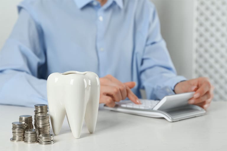A man using a calculator next to a model tooth and a stack of silver coins