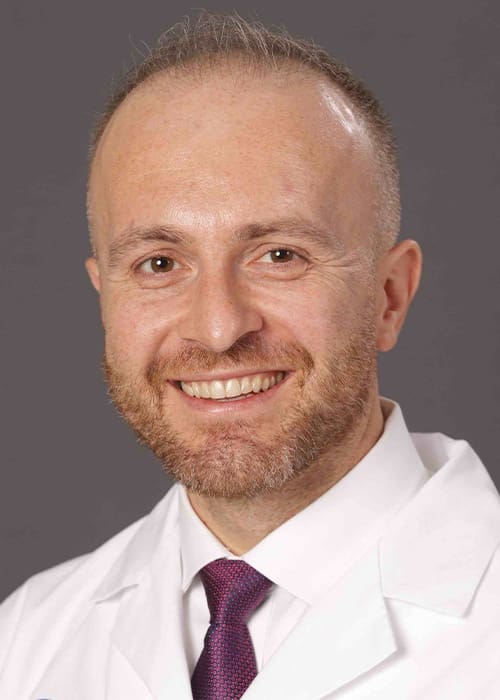 Profile image of Dr. Konstantinos Vazouras smiling with a white lab coat and purple tie.