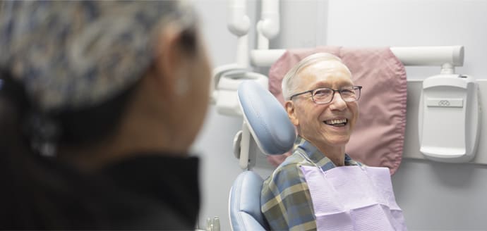 An elderly man in glasses smiles warmly while seated in a dental chair, wearing a plaid shirt and a bib, with a dentist blurred in the foreground.