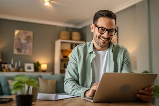 Man with glasses smiling while working on laptop at home.