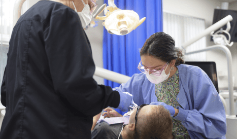 Two dental professionals in masks and scrubs attend to a patient in a dental chair. Overhead light illuminates the scene. Mood is clinical and focused.