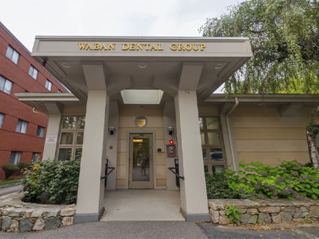 Front view of Waban Dental Group, a single-story building with a neutral facade and greenery. It has a welcoming, professional dental office entrance.
