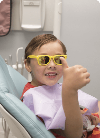 A young boy with yellow sunglasses sits in a dentist chair at Waban Dental Group, smiling and relaxed.