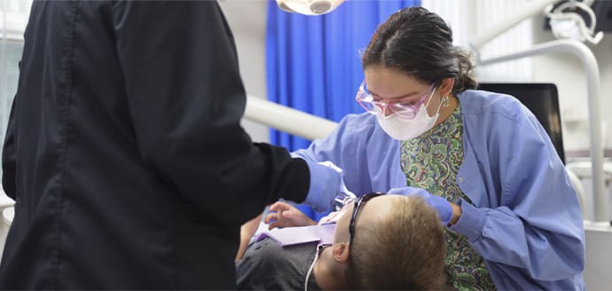 Two dental professionals in masks and scrubs attend to a patient in a dental chair. Overhead light illuminates the scene. Mood is clinical and focused.