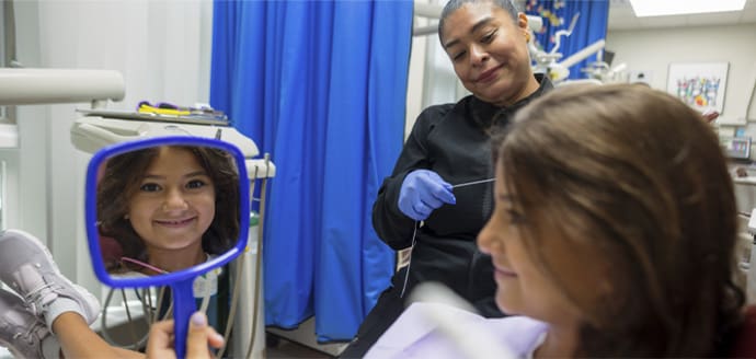 A young girl at the dentist's office smiles at her reflection in a hand mirror. The dentist, wearing gloves, stands nearby in a friendly atmosphere.