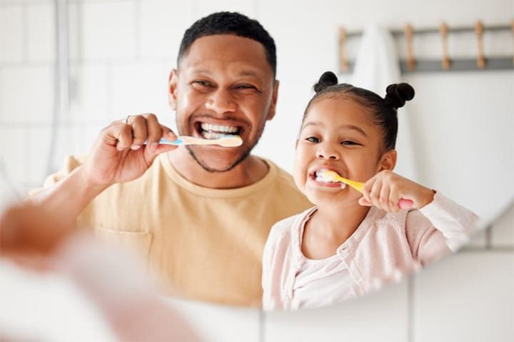 A man and a young girl brushing their teeth together, promoting dental hygiene and family bonding.